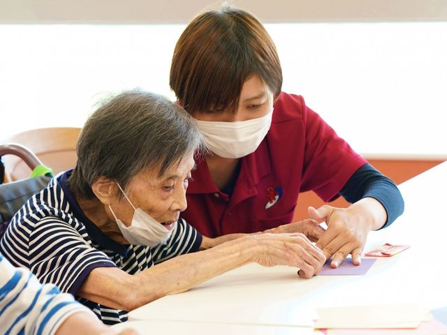 介護職員と利用者の活動 スタッフイメージ スタッフが利用者と一緒にテーブルでの作業をサポートしています。和やかな雰囲気が感じられます。