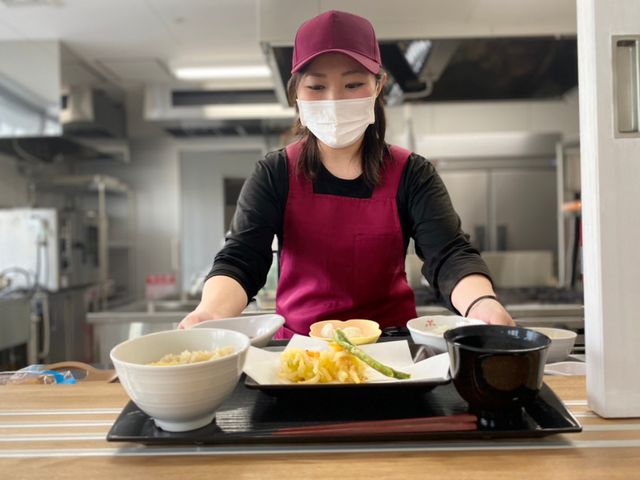 料理を配膳するスタッフ 食事例 丁寧な手作りの食事を提供しており、笑顔で配膳するスタッフの様子が活気に満ちています。ここでは心温まるサービスを実感いただけます。