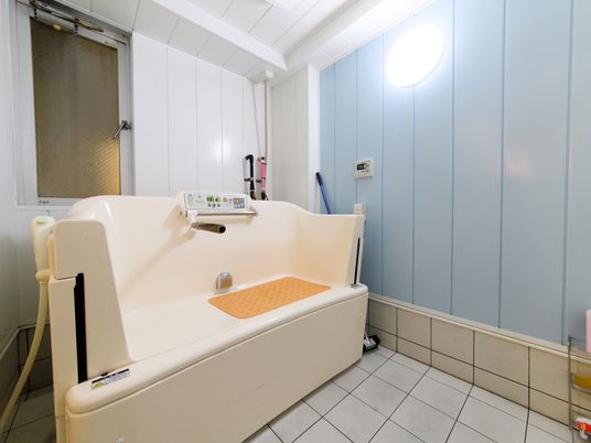 施設の写真 浴室の床はタイルになっている。窓側の壁は白色だが、ライトのついている壁は水色である。浴槽にオレンジのすべり止めマットが敷いてある。