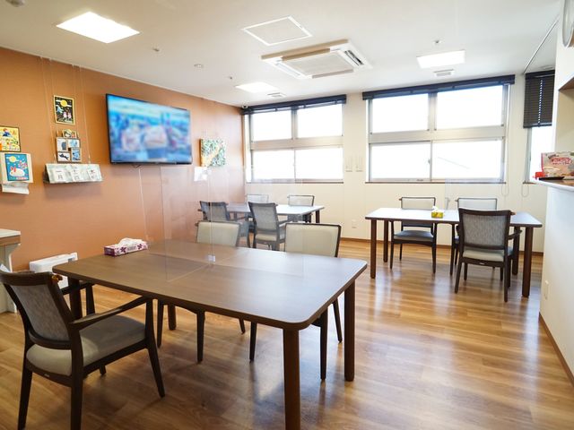 共有スペースの居心地の良さ 共有スペース 温かみのある壁紙と木目調の床が印象的なダイニングルームです。壁掛けのテレビや各種情報が配置され、快適な休息空間を提供しています。
