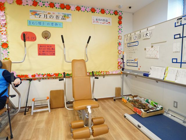 リハビリの設備充実の部屋 機能訓練室 座席に座る利用者の傍らには、トレーニングに专用された様々な器具が整えられた機能訓練の空間です。周囲の壁には目標や日程が掲示され、活動的な雰囲気が漂っています。