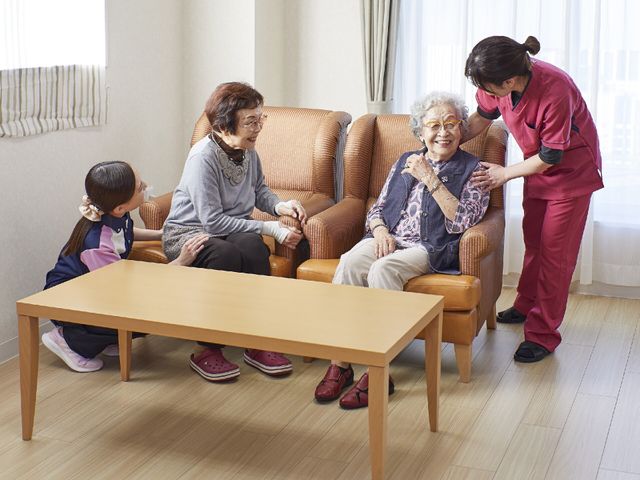 居室での患者様とスタッフ サポート ゆったりした椅子に座り、スタッフと笑顔で会話する患者様の和やかな様子です。明るい室内でのひとときをご覧ください。