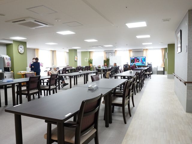 広々とした明るい食堂 共有スペース ゆったりと配置されたテーブルが並ぶ食堂では、利用者が心地よい時間を過ごしています。スタッフの方々も笑顔で対応しており、明るい職場環境が伺えます。