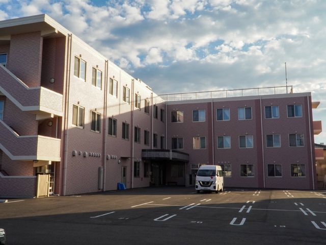 ピンクの建物の外観 外観 清々しい朝の光に包まれた、ピンク色の落ち着いた雰囲気の建物です。広々とした駐車場には、移動の便を図るための車両も確認できます。