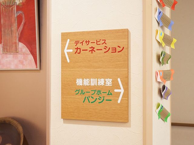 機能訓練室の案内板 廊下   木目調の案内板があり、デイサービスや機能訓練室の方向がわかりやすく示されています。廊下の壁には飾りがあり、明るく親しみやすい雰囲気です。