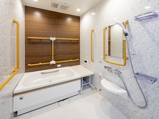 施設の写真 ベージュの手すりがたくさん付いた浴室には一人用の浴槽が設置され、その横にはベンチが置かれている。