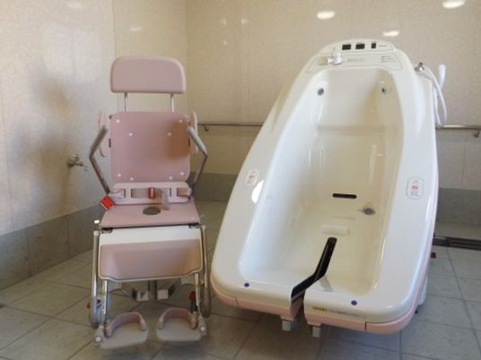 浴室用の車椅子が用意された機械浴室である。清潔感のある空間で、壁に銀色の手すりが取り付けられている。