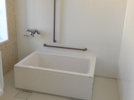 白を基調としたシンプルな浴槽が設置されている。壁には、茶色の手すりが２つと蛇口が取り付けられている。