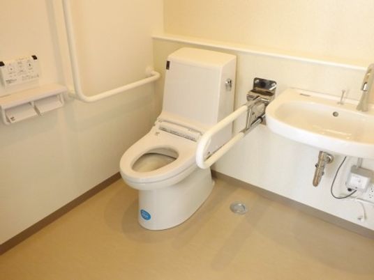 洗面台が備え付けられた個室トイレである。壁にＬ字型の手すりや操作パネル、ペーパーホルダーが設置されている。