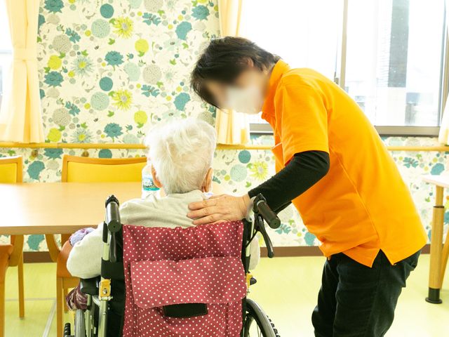 ケアスタッフと利用者様の背中 スタッフ 明るく開放的な空間で、優しい手つきのスタッフが利用者様の肩を支えています。温もりのある接客が行われている様子が伝わってきます。
