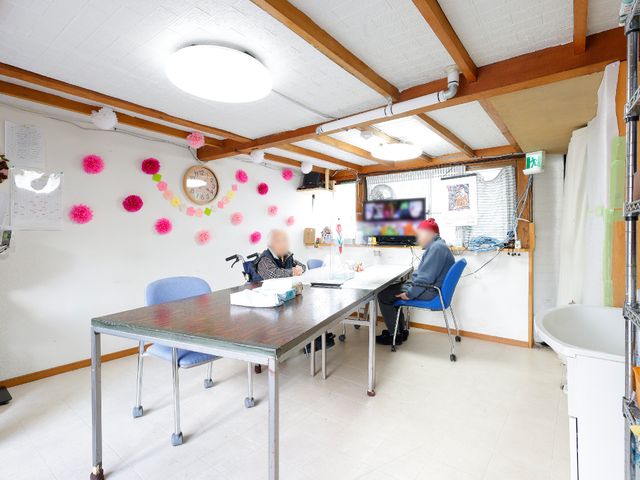 多目的利用可能な居室 共有スペース 落ち着いた雰囲気の広々とした部屋で、一人の方がテーブルに向かい静かに活動されています。このような空間は、日々の業務にも前向きな気持ちを与えてくれるでしょう。