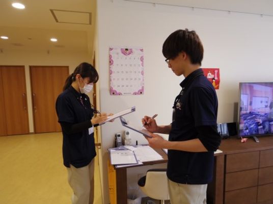 紺のシャツを着た２人の職員が、書類を見ながら情報共有をしている。施設では、24時間体制で職員が入居者様のサポートを行っている。