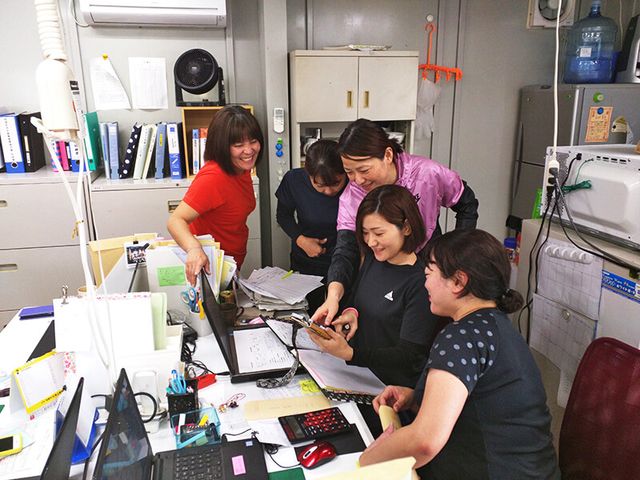 スタッフが相談中の事務室 スタッフイメージ スタッフが和やかに業務内容を相談しています。チームワークを大切にする職場環境です。