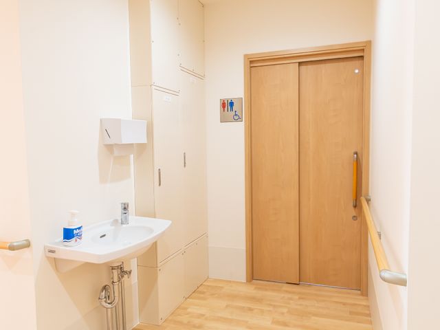 手洗い場とトイレの扉 洗面台 手洗い場にはせっけんが置かれ、清潔を保つ工夫がされています。トイレの扉には、車椅子を示すマークがあり、バリアフリーへの配慮が見受けられます。