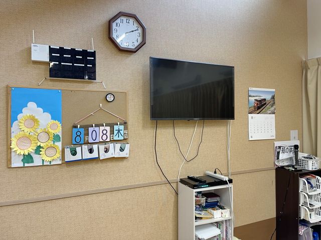 時計とテレビのある居室 共有スペース 壁に掛かる時計と、向かいに設置された大画面テレビが配置された居室です。壁面には季節感を感じさせる装飾もあります。