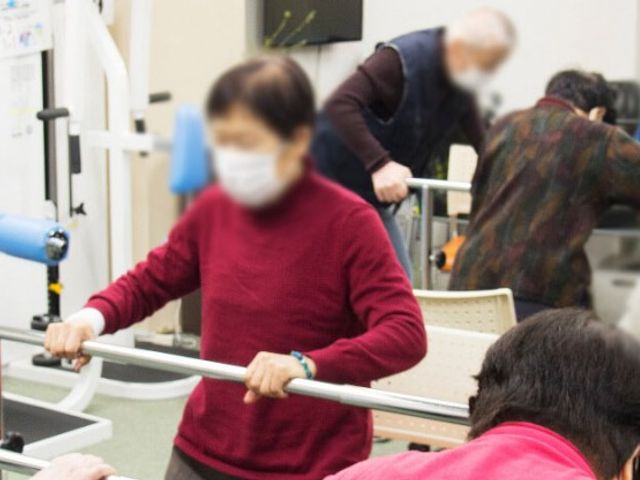 利用者が運動する室内 機能訓練室 明るい空間で、多くの利用者が運動器具を使って身体を動かしています。和やかな雰囲気で、会話も弾む環境です。
