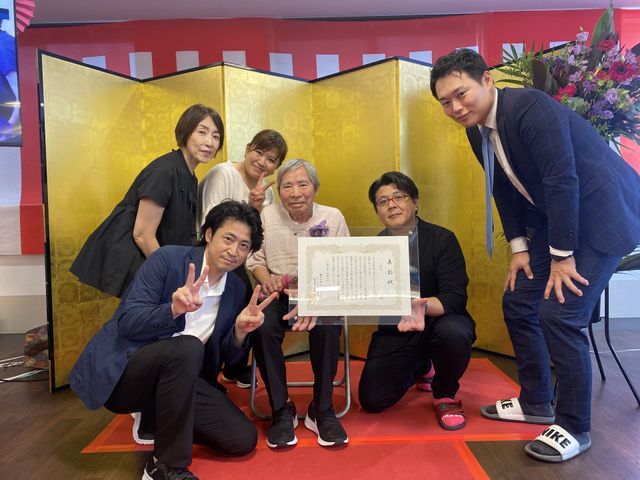 式典を祝う人々 多目的室 和やかな雰囲気の中、表彰式が行われています。受賞者を囲む職員の笑顔が輝いています。