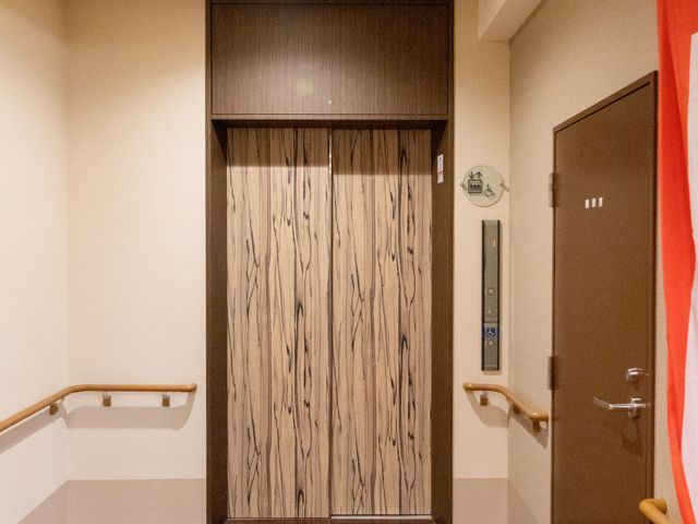 木目扉のエレベーター前 エレベーター   木目調の扉が落ち着いた印象のエレベーター前です。手すりがあり、移動しやすい配慮が見られます。