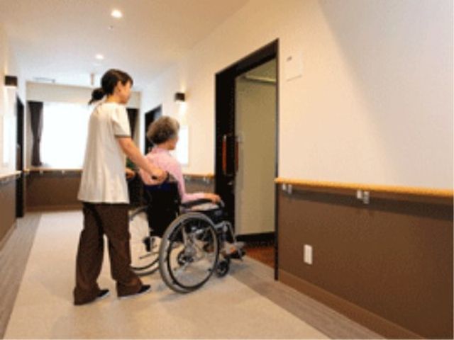 車椅子利用者とスタッフ廊下 出入口 ゆったりとした廊下で、職員が笑顔で車椅子を押しています。壁には手すりが設置され、安全に配慮されたつくりになっております。