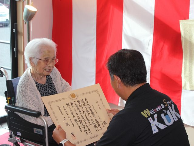 表彰式の様子 スタッフイメージ 入所者様が表彰を受ける和やかな場面です。スタッフとの交流が温かく、心の通う瞬間です。