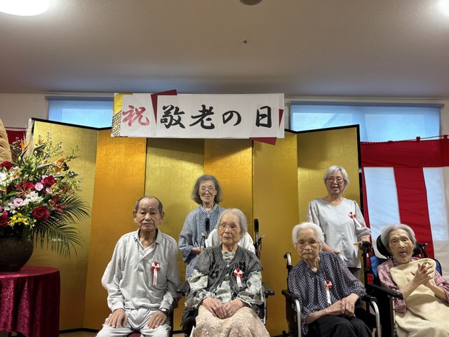 敬老会に参加する入居者 リビング 敬老の日を祝う会にて、入居者の皆さんが楽しそうに記念撮影をしています。和やかな雰囲気です。