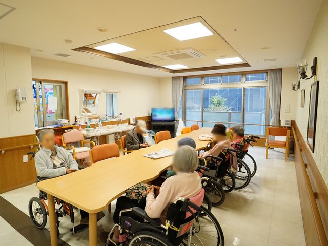 温かみのある居室空間 共有スペース 広々とした明るい居室で、入居者の方々が談笑されています。椅子に座り、快適な時間を過ごされています。