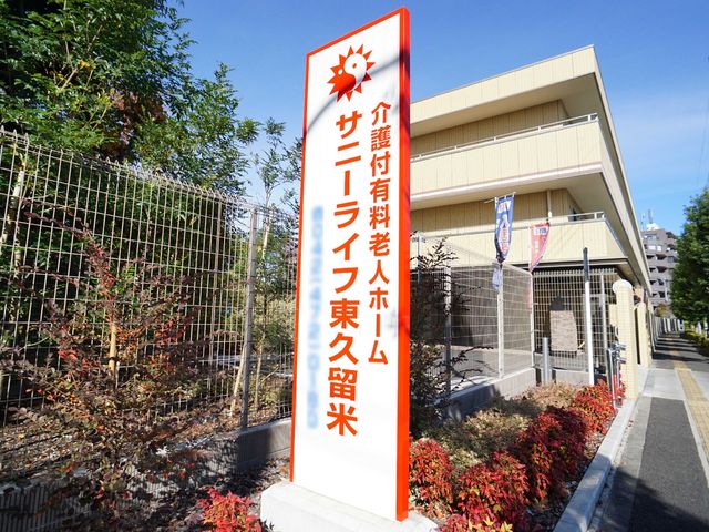 サインボードのある景観 ロゴ 清潔感のある明るい建物の前に立つ案内看板です。日差しに包まれ、落ち着いた雰囲気が感じられます。