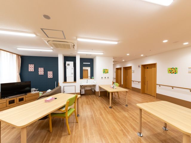 明るい共有スペース リビング   木目調の床とやわらかな照明で、落ち着いた雰囲気の共有スペースです。テレビやテーブルがあり、入居者さまが過ごしやすい環境に見えます。