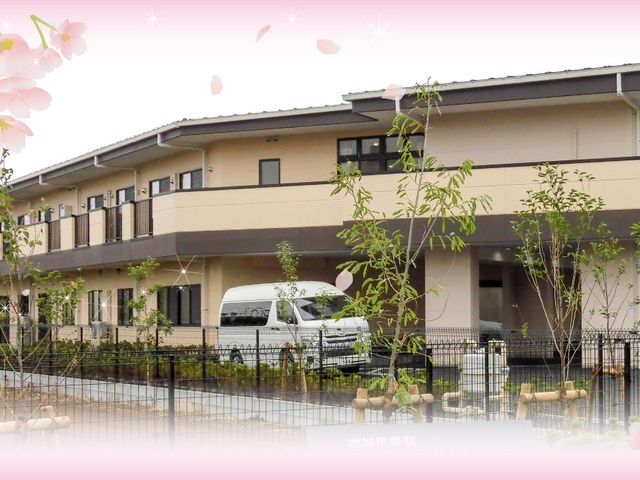 施設外観 外観   落ち着いた色合いの2階建てで、前面に植栽が整えられています。送迎車が停めやすそうな駐車スペースも見えます。