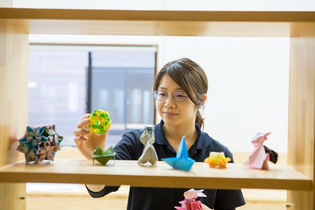 スタッフの活動風景 スタッフイメージ スタッフが折り紙を手に取りながら笑顔で様々な色と形の作品を見ています。手先を使った活動が日々提供されている様子がうかがえます。