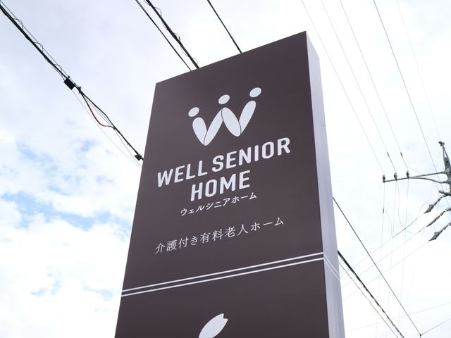 高齢者向け施設の外の看板 看板 青と白の空の下、施設のシンボルと名前が記された看板が目を引きます。訪れる方々への心地よいお出迎えを象徴しています。