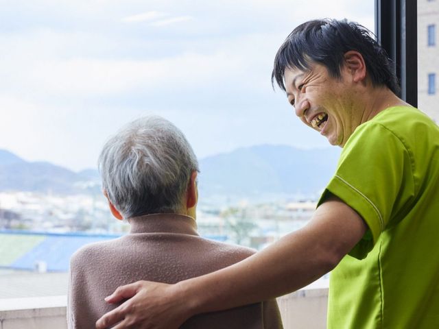 笑顔で会話するスタッフと利用者 スタッフイメージ 笑顔溢れるスタッフが利用者様の肩に優しく手を置き、心温まる会話を交わしています。
