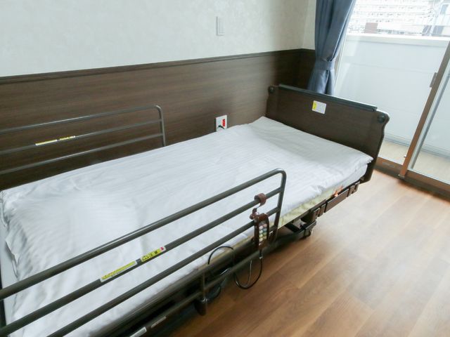 居室のベッドと窓 ベッド きれいに整えられた居室には、窓から自然光が差し込み、明るく穏やかな雰囲気を感じさせます。手すり付きのベッドは安全性を考慮して設計されており、快適な生活空間が確保されています。