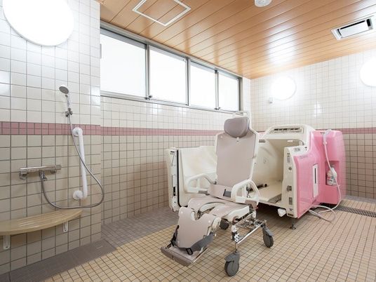 白とピンクの機械浴を設置したタイル張りの浴室。座ったまま入浴できるタイプの機械で、シャワーも付いている。