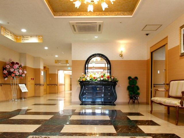 エントランスの花と椅子 エントランスホール 花々で飾られたアーチ型の入口が印象的なエントランスです。落ち着いた色合いの椅子が設けられ、待ち合わせにも適しています。