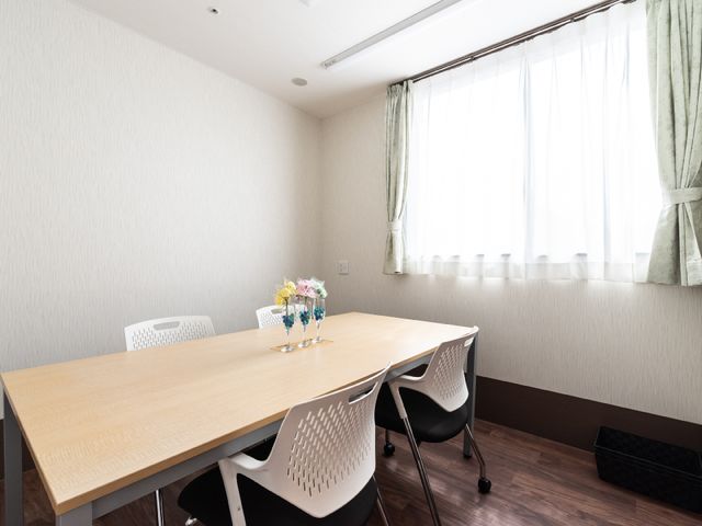 明るい居室のテーブルとイス 相談室 落ち着いた色合いの居室です。天窓から自然光が差し込み、テーブルに花が飾られています。