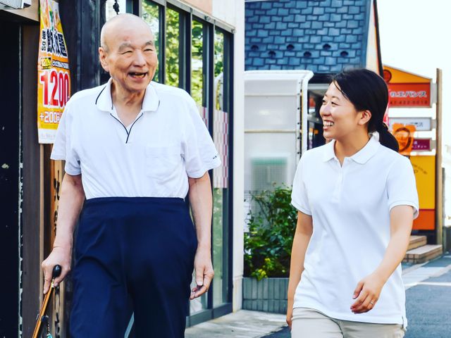 散歩する高齢者と介護者 あたたかな笑顔で高齢者の方との散歩を支援するスタッフの姿が見られます。明るく積極的な関わりを大切にしています。
