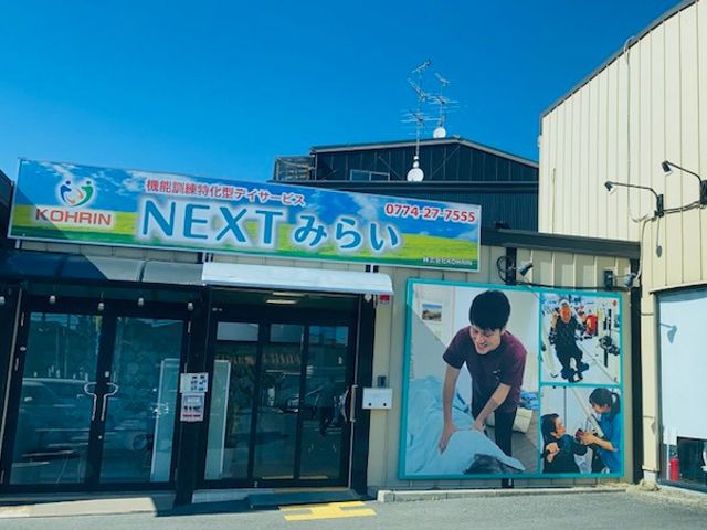 看板のある建物外観 外観 明るい日差しの中、笑顔のスタッフがケア活動をしている様子が見られます。優しさと活気を感じる環境です。