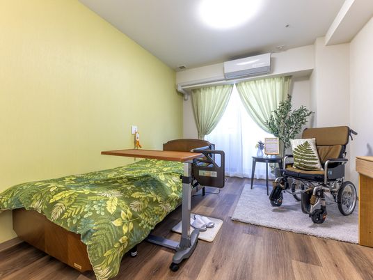 施設の写真 寝具付きのベッドサイドには緊急通報装置があり、車いすも置かれているフローリングの部屋にはエアコンもついている。