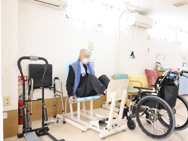 介護用機器と椅子の部屋 機能訓練室 広々とした空間には、機能訓練に使われる様々な器具が整えられ、利用者様のリハビリをサポートしています。明るく清潔感のある環境で、毎日の訓練に励むことができます。