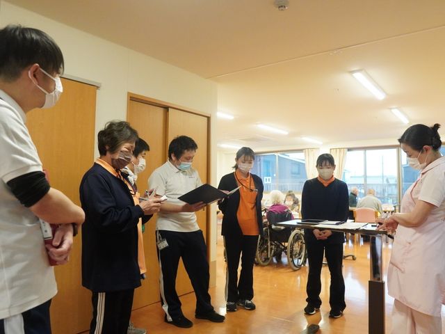 スタッフミーティング風景 ラウンジ スタッフが円になり、日常業務の確認を行っています。背後には自然光が差し込む明るい共有スペースが広がっています。