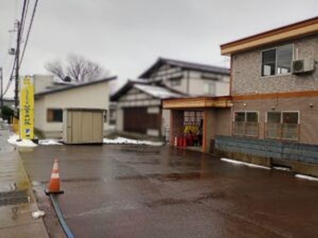 建物前の駐車場 外観   雨で濡れた駐車場と建物の外まわりが見えます。出入口付近まで車で近づきやすそうなつくりです。