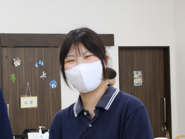 マスク姿の女性と室内 内観   マスクを着けた女性が笑顔で立っており、明るい室内の雰囲気が伝わります。就業時のやわらかな印象をイメージしやすい場面です。