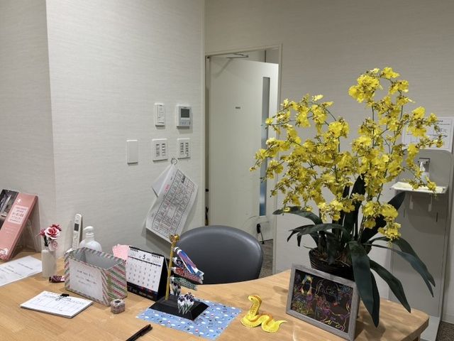 フロントの受付デスク フロント   木目調の受付デスクに花や小物が並び、落ち着いた印象です。出入口の先に続く空間も見え、来訪対応のしやすさがうかがえます。