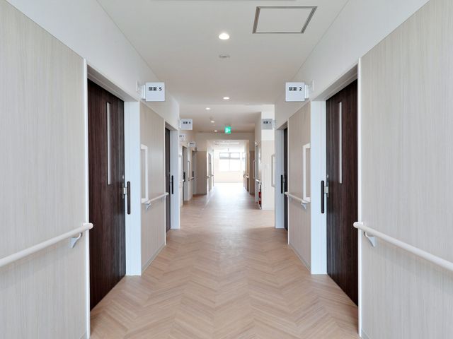 白と茶の配色の廊下 廊下 手すり付きで広々とした廊下は明るく、歩行にも安心感をもたらしています。扉沿いには番号が見やすく配されており、ご自身の居室へのアクセスも容易です。