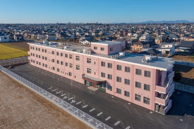 ピンクの外壁の建物外観 外観 広々とした駐車スペースが整えられた新しい建物です。明るい色の外壁が印象的で、快適な環境での就業が期待できます。