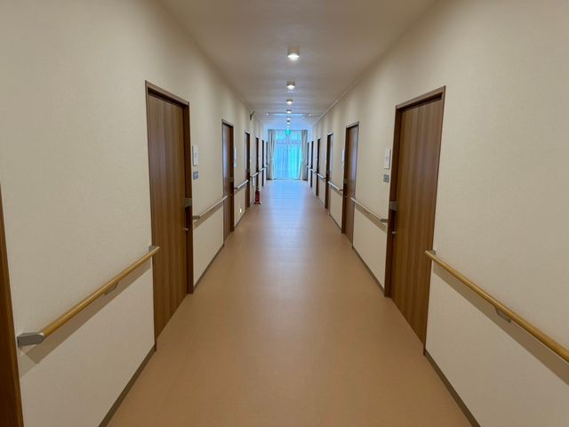 手すり付き廊下 廊下   両側に手すりが設けられた、まっすぐな廊下です。奥に自然光が入り、移動しやすい落ち着いた環境です。