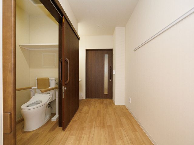 バリアフリートイレ居室 トイレ 手すり付きのトイレが備わった居室で、安心して生活を送ることができます。木目調の床と壁が温かみのある空間を演出しています。