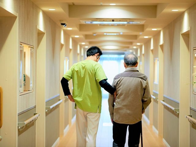 支援を受ける利用者と職員 日常風景 職員が手を添えながら歩く安全な廊下です。手すりが整備され、ゆったりと心地よい時間を過ごせる設計になっています。
