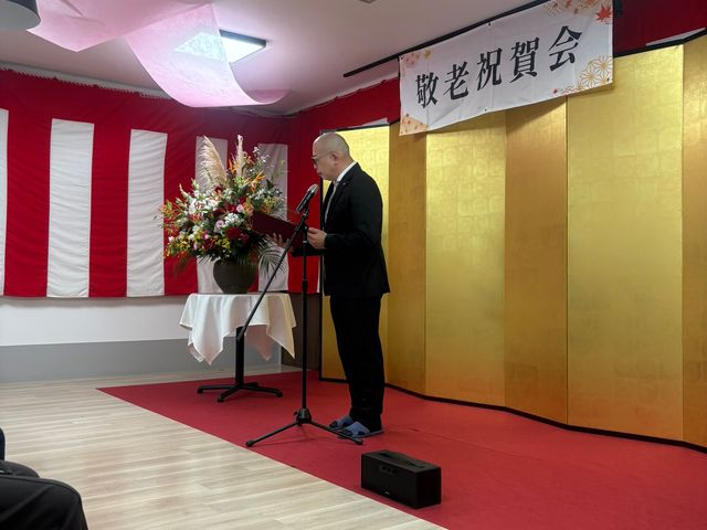 敬老会の式典風景 多目的室 敬老祝賀会の壇上でスピーチが行われています。参加者が集まり、華やかな雰囲気です。